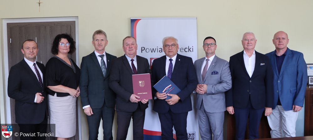 Grupa ośmiu przedstawicieli Powiatu Tureckiego i Poznańskiej Akademii Medycznej pozuje do zdjęcia po podpisaniu listu intencyjnego. W centrum Starosta Jan Smak i Rektor Włodzimierz Usarek trzymają dokumenty. W tle baner z logo Powiatu Tureckiego.