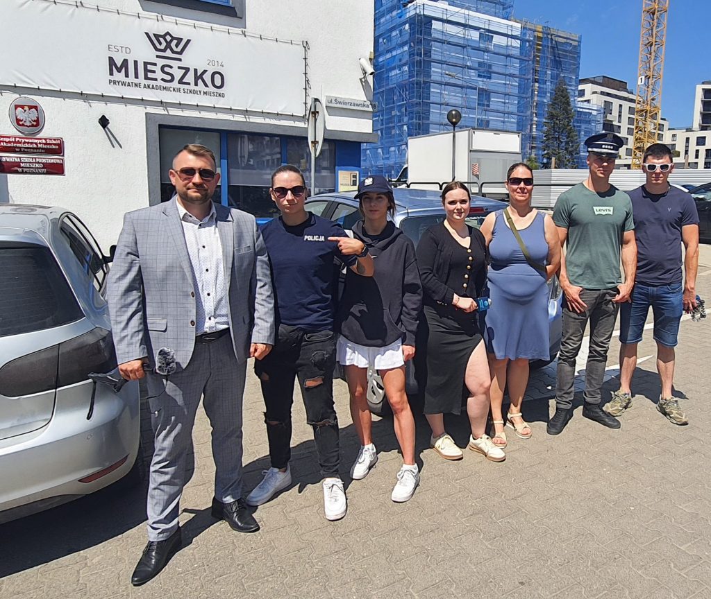 Na zdjęciu grupa siedmiu osób stoi na parkingu przed budynkiem Prywatnych Akademickich Szkół Mieszko. Część osób ma na sobie elementy stroju policyjnego, inni są ubrani swobodnie lub elegancko. W tle widoczny jest szyld szkoły MIESZKO